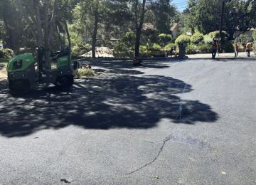 Pothole repair crew patching asphalt road beside a green paving machine in a tree-lined park area